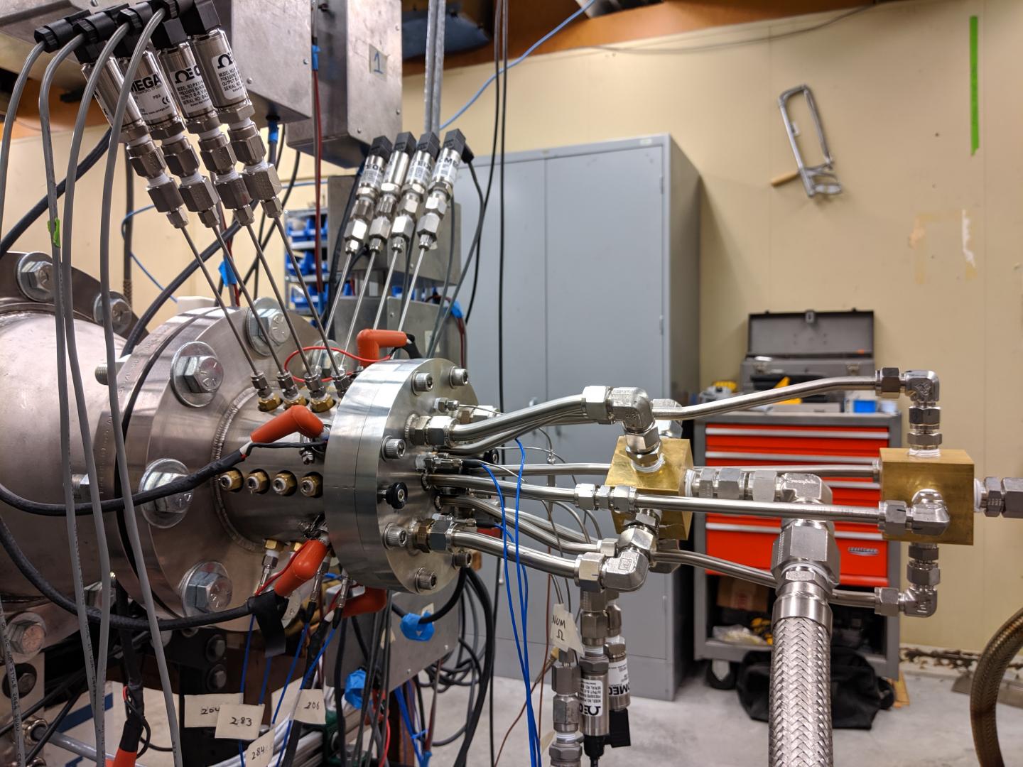 CAPTION The researchers first developed an experimental rotating detonation engine (shown here) where they could control different parameters, such as the size of the gap between the cylinders. The feed lines (right) direct the propellant flow into the engine. On the inside, there is another cylinder concentric to the outside piece. Sensors sticking out of the top of the engine (left) measure pressure along the length of the cylinder. The camera would be on the left-hand side, looking from the back end of the engine.  CREDIT James Koch/University of Washington