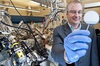 Purdue professor Michael Manfra holds a gallium-arsenide wafer as he stands next to the high-mobility gallium-arsenide molecular beam epitaxy system, or MBE, at the Birck Nanotechnology Center. Manfra leads a team of Purdue researchers that create ultrapure semiconductor heterostructures to study electrons in correlated states. (Purdue University photo/Andrew Hancock) 